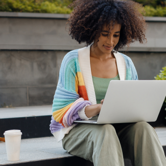 Young woman with natural hair and cozy cardigan sits on steps outside while working on her laptop.
